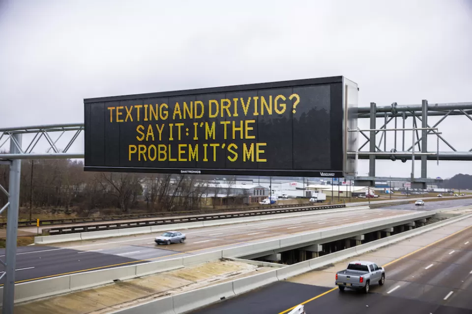Digital highway sign above six-lane highway that says, "Texting and Driving? Say It: I'm the Problem It's Me."